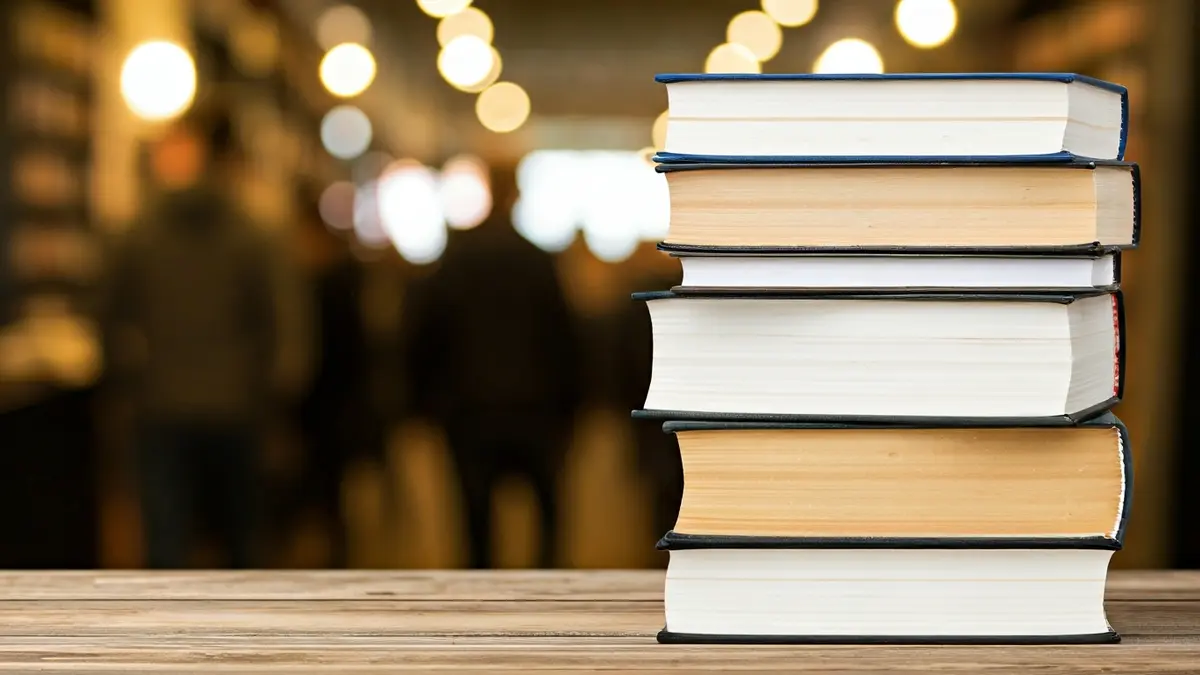 Imatge genèrica de llibres apilats en una taula de fusta, amb un fons desenfocat d'una biblioteca.