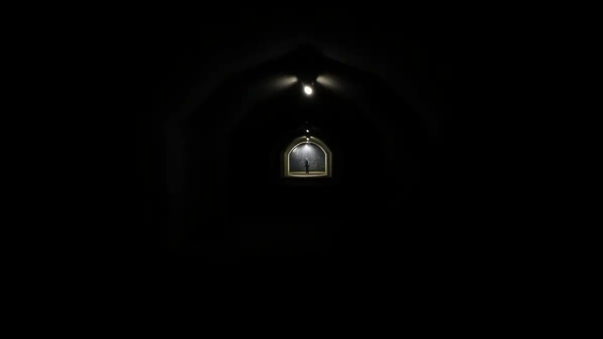 Imagen de un túnel subterráneo oscuro y vacío con paredes de hormigón y un techo bajo, iluminado por una única fuente de luz tenue en la distancia, creando sombras largas y una sensación de misterio.