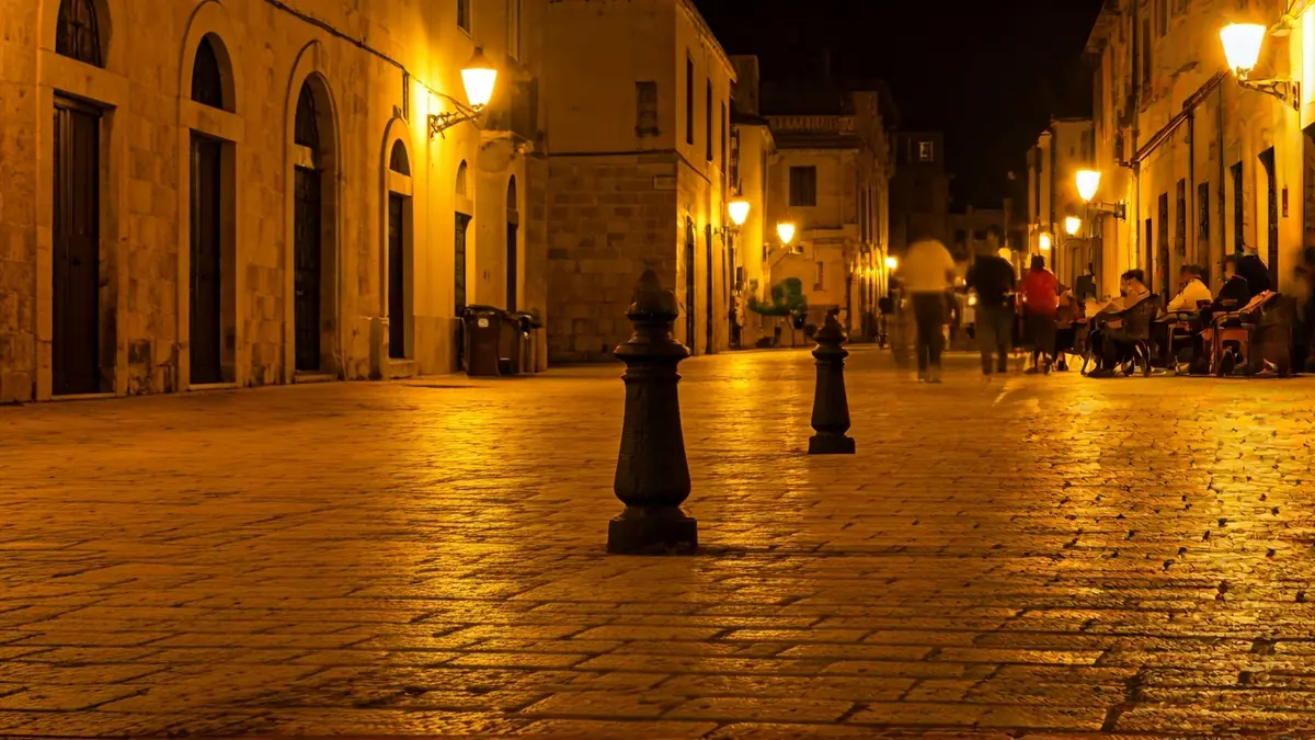 Generic image of a Mediterranean square at night, with streetlights and blurred figures in the background.