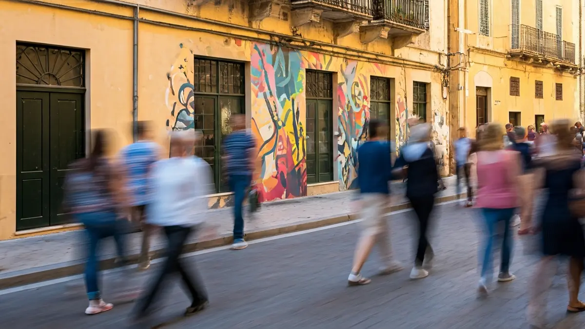 Imagen de un mural artístico en la fachada de un edificio en una ciudad mediterránea, con figuras borrosas de personas y una atmósfera festiva.