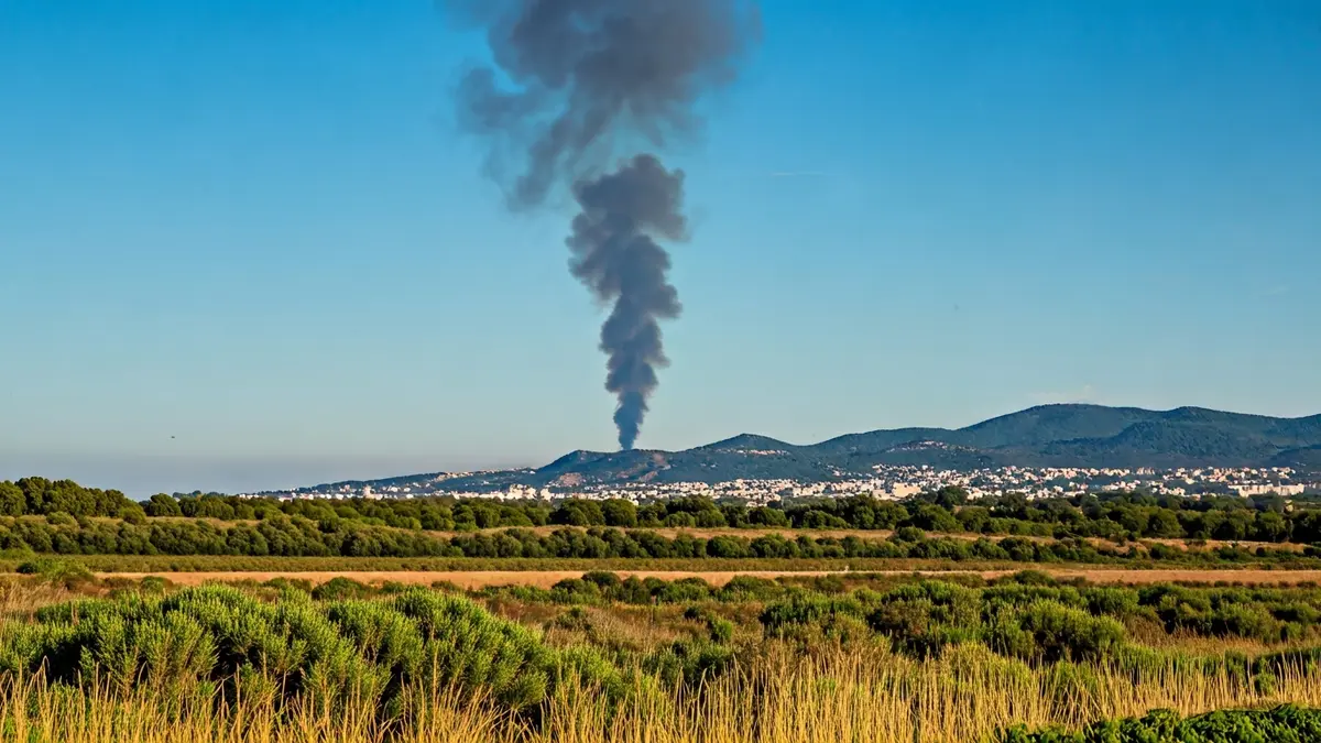 Imatge genèrica d'una columna de fum gris elevant-se sobre un paisatge mediterrani amb vegetació i cel blau.