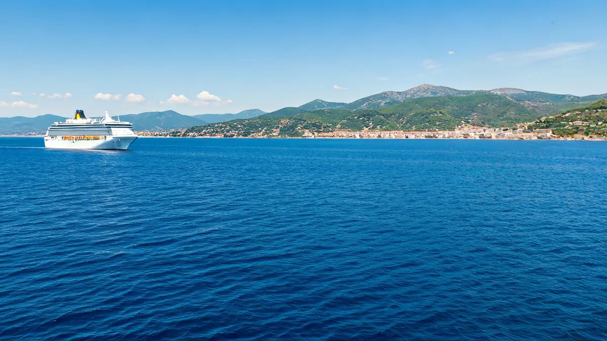 Imatge genèrica d'un creuer navegant per aigües blaves amb la costa mediterrània al fons.