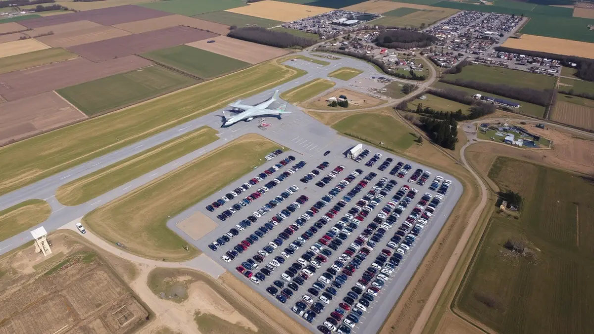 Imagen aérea de una gran explanada de aparcamiento llena de vehículos cerca de un aeropuerto, rodeada de paisaje rural y algunas casas dispersas.