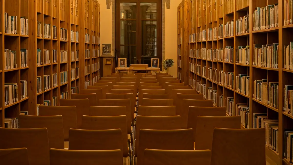 Image of a warm-lit exhibition hall or library with wooden bookshelves and a podium with a microphone, in a Mediterranean-style building.