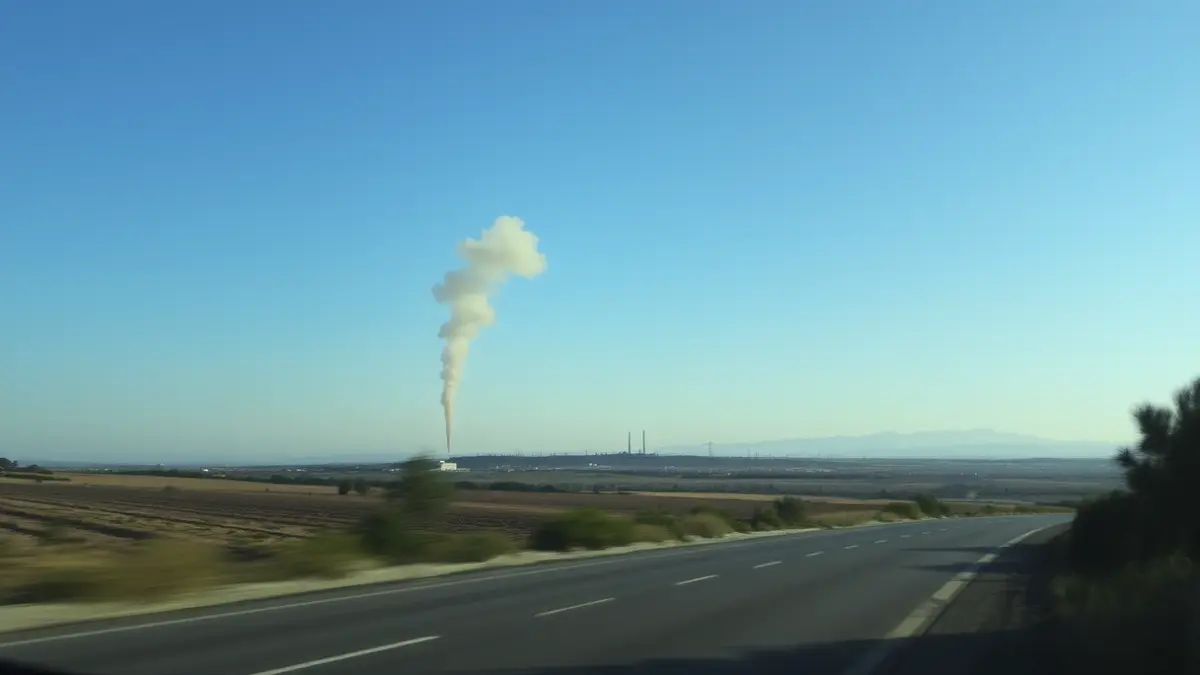 Imagen genérica de una columna de humo gris elevándose sobre una zona industrial, con una carretera en primer plano y un paisaje mediterráneo.